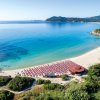 Spiaggia con ombrelloni rossi su sabbia chiara e mare calmo, vista da una collina.
